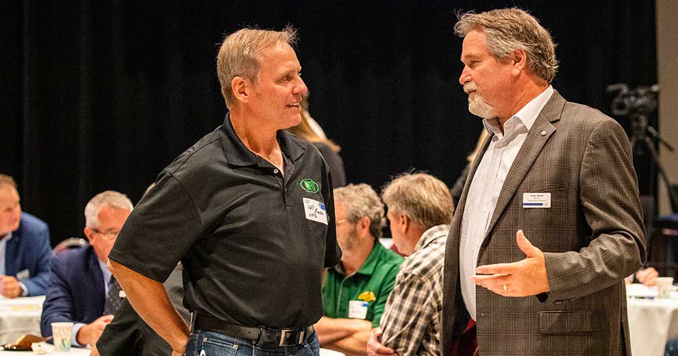 Two men talking at meeting with group of people at a table in background