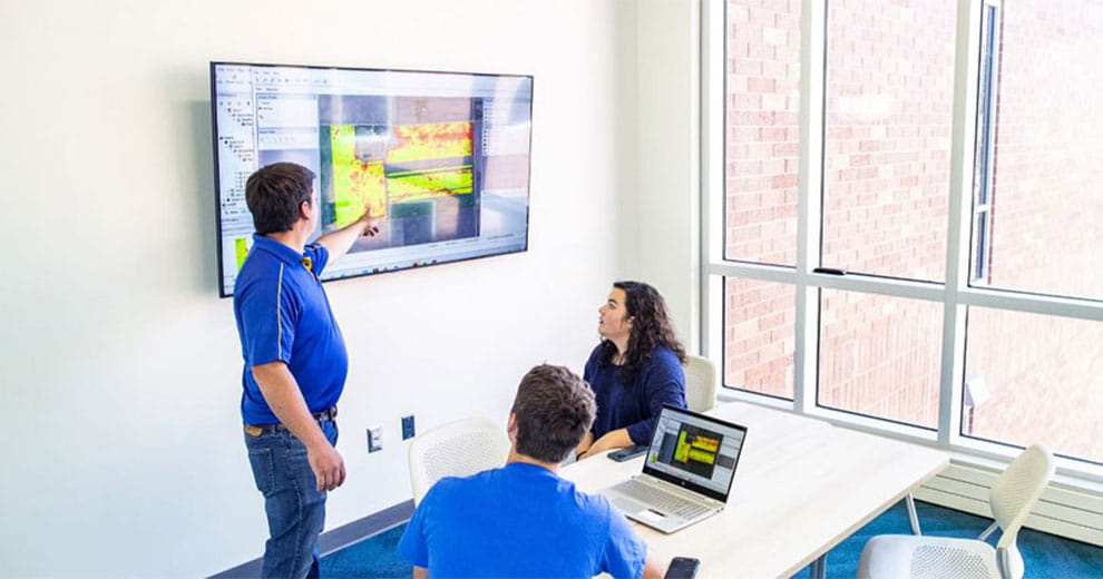 Man standing and pointing at graph on a TV screen while seated man and woman look to where he's pointing