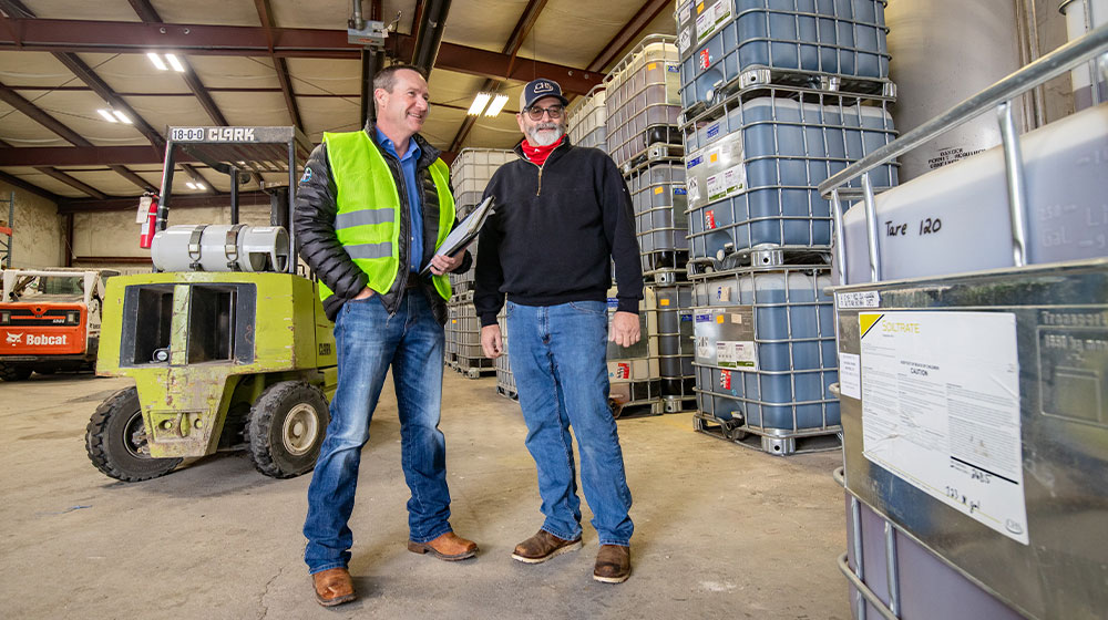 Two men standing in a warehouse near farm chemicals.