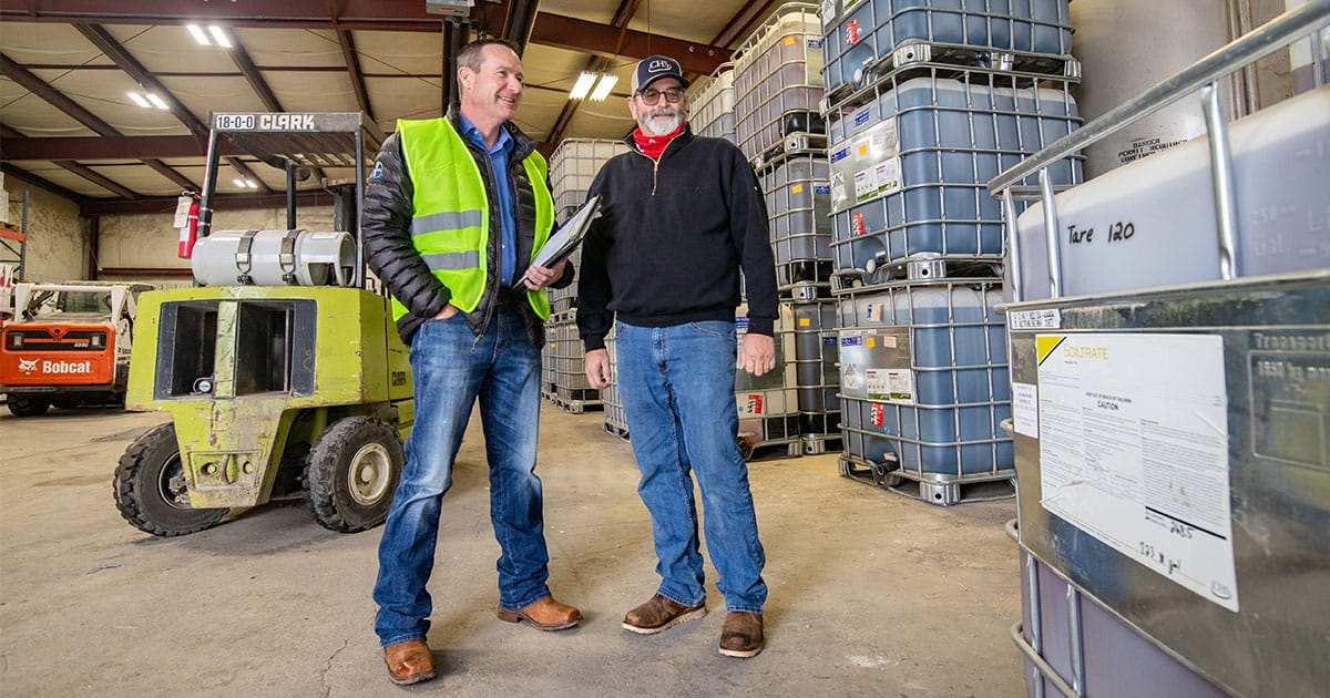 Two men standing in a warehouse near farm chemicals.