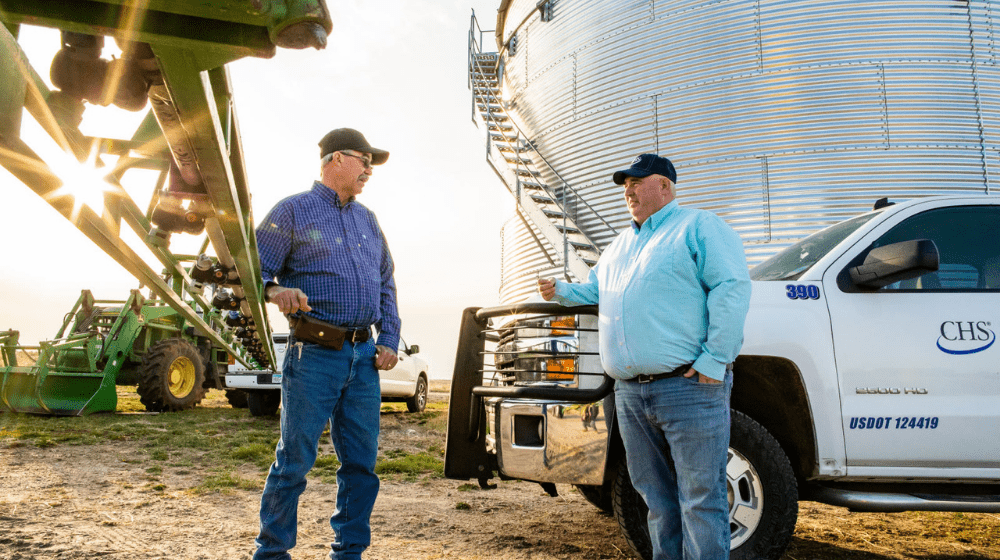 Two men standing outside near a sprayer and grain bin.
