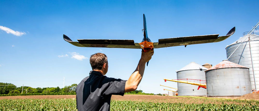A man holding a drone stands in a corn field behind some grain bins.
