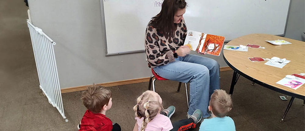 Woman on chair reads book to group of children sitting on floor