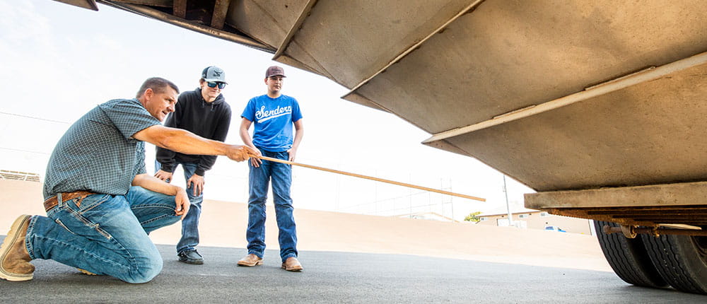 An instructor and two high-school students look at the underside of a large truck