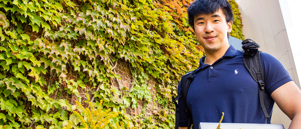 Student holding laptop standing in front of an ivy-covered wall