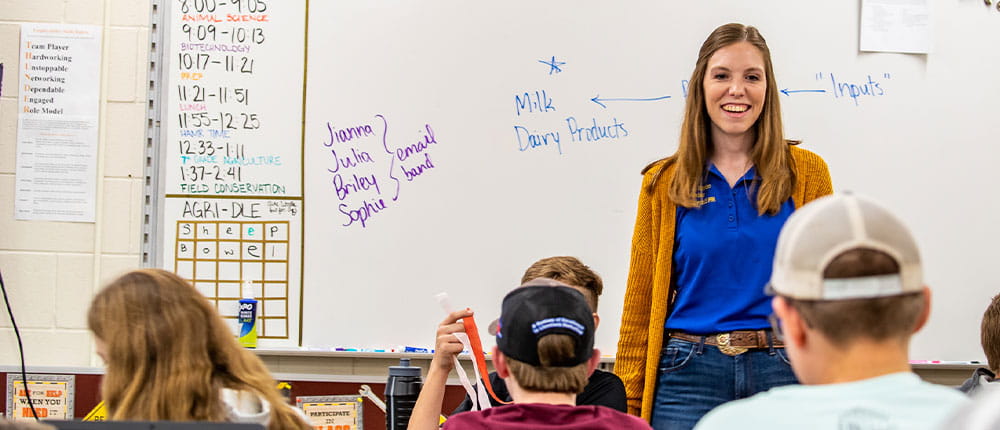 A teacher in front of a classroom with a whiteboard behind her