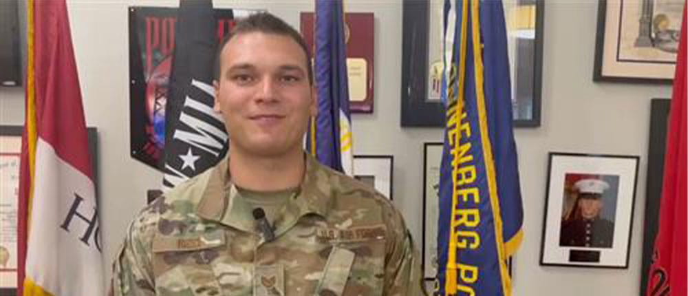 Man in military fatigues standing in front of flags and framed photos