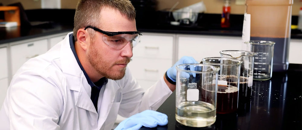 Man wearing safety glasses, blue disposable safety gloves and white lab jacket bends down to countertop to look closely at liquids in test tubes.