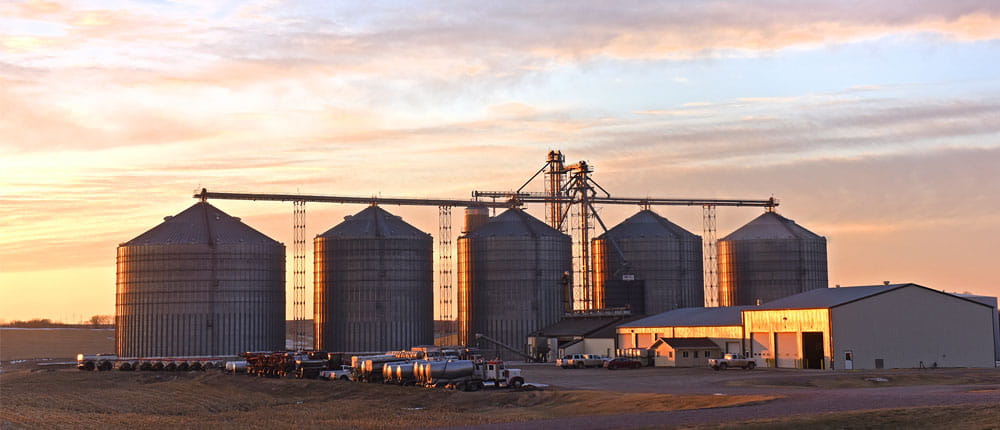 A sunset is the backdrop of five grain bins on a farm.