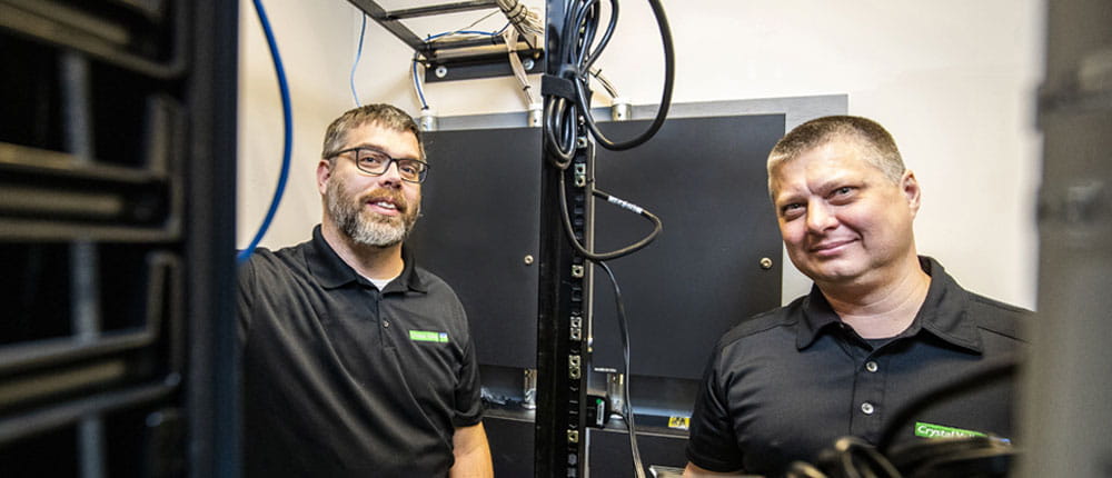 Two men stand in front of computer servers.