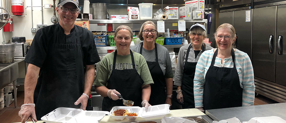 Five people in an industrial kitchen standing behind a counter with food packaged for takeout