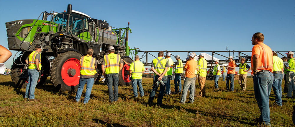 A group of people wearing bright yellow safety vests stands next to a sprayer