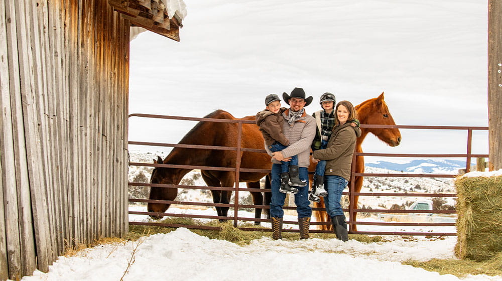 Family stands by barn and horses with a snowy mountain backdrop