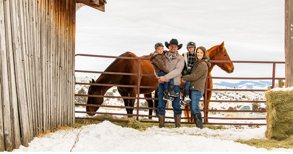 Family stands by barn and horses with a snowy mountain backdrop