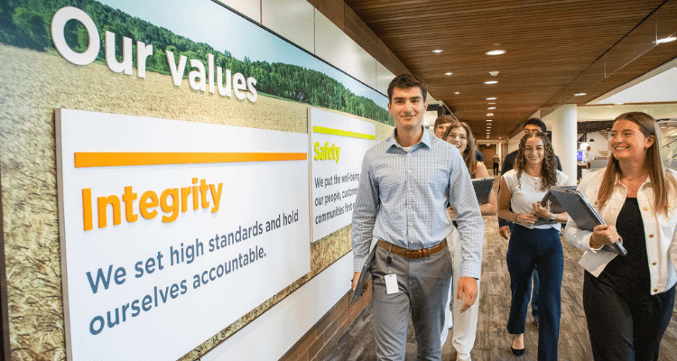 Group of interns walk down an office hallway beside a wall display highlighting CHS values, including integrity and safety